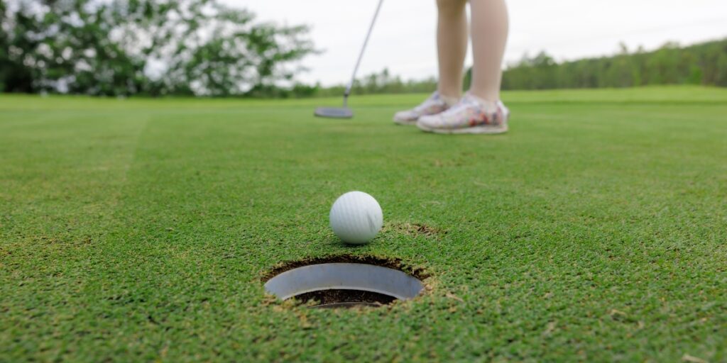 golf ball in front of the hole with a golfer with putter in the background